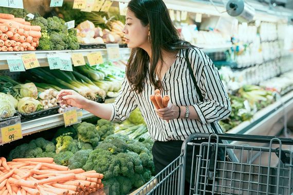 A woman holding some carrots by a cart, shopping for produce in a grocery store